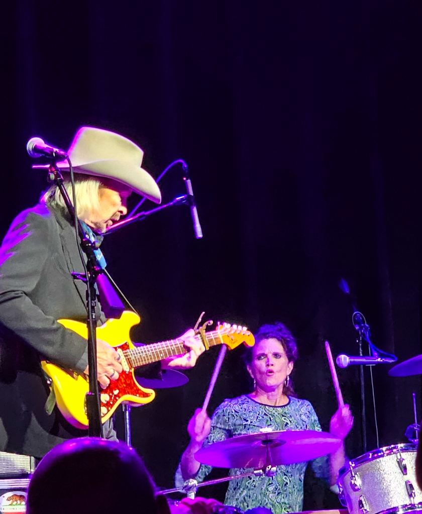 Light skinned man in dark blazer and cowboy hat playing guitar, light skinned woman playing drums
