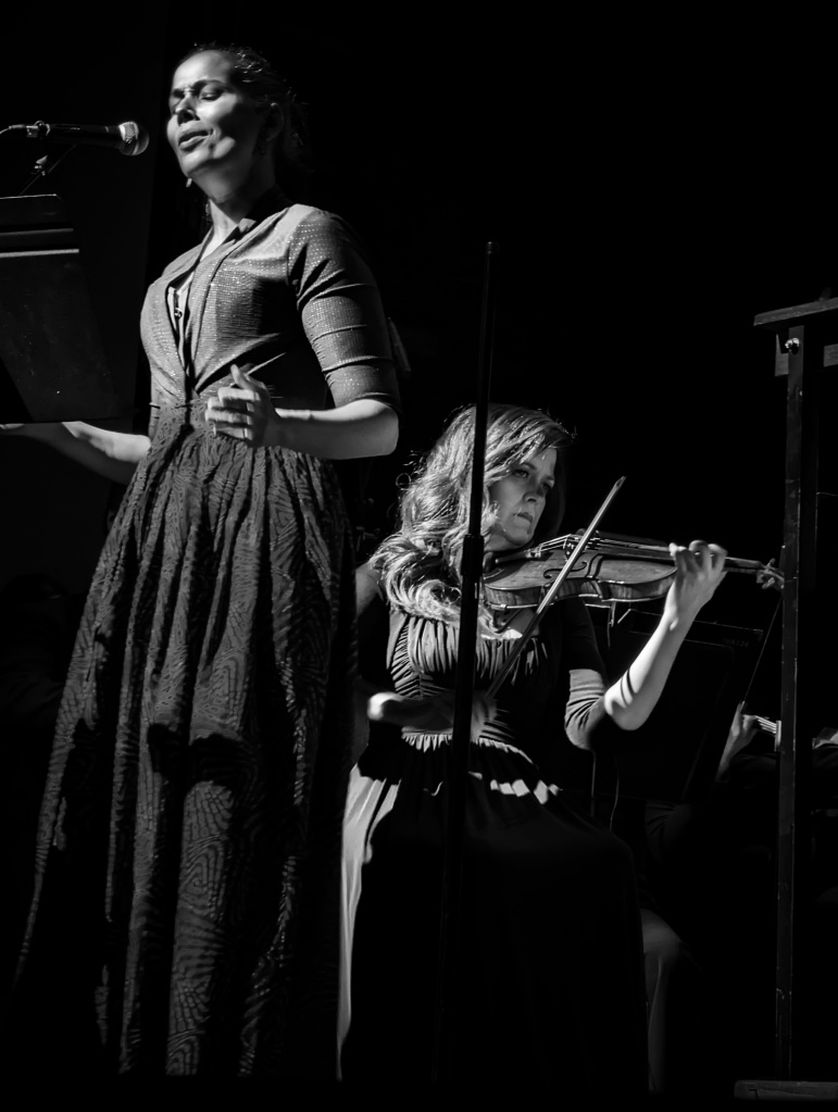 Black and white photo, dark skin woman singing, light skinned woman sitting and playing violin