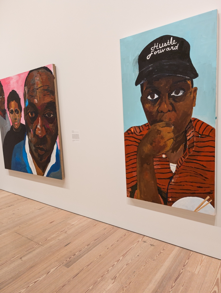 Two large photos on the wall of the Whitney, one three black people arrayed (from left to right) a man in the background, a woman in the middleground, and a man in the foreground. The other, a man wearing a hat that says Hustle Forward and an orange and black striped shirt with a bowl with chopsticks in front of him